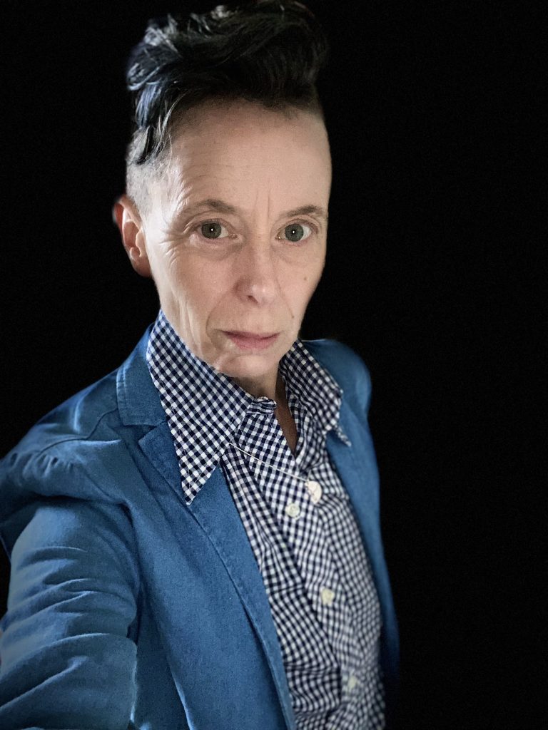 A white person wearing a gingham top under a bluebird blue jacket holds up an arm to take a selfie a little off to the side. Their dark hair is shaved on the sides and slicked back on top. They might have a tiny smirk. Or maybe it's a glower. They are gaunt and the camera is far enough away that it's almost hard to see their many wrinkles.
